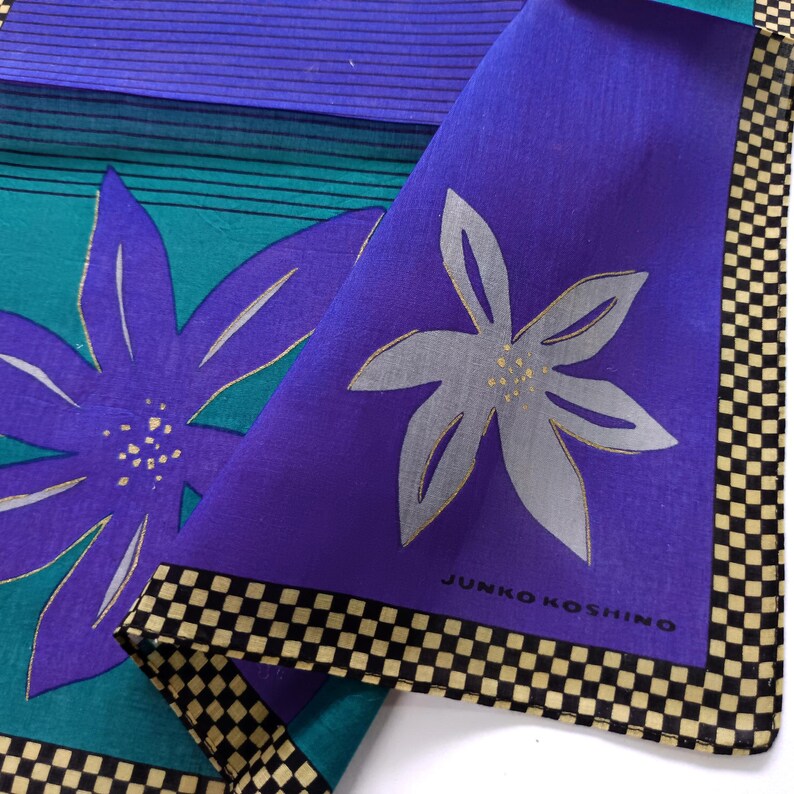 May include: A folded square textile with a floral design. The textile features a large purple flower with silver accents on a teal background. A second section is purple with a silver flower. The border is black and tan checkered. The text "JUNKO KOSHINO" is visible.