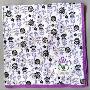 May include: White handkerchief with a floral pattern in black, gold, and purple. The border is purple. The handkerchief has the embroidered text "ANNA SUI" and a floral design.