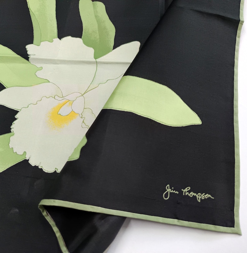 Puede incluir: Pa&ntilde;uelo de seda negro con un gran dise&ntilde;o de orqu&iacute;dea blanca con detalles en amarillo y verde. El pa&ntilde;uelo tiene un borde verde claro y la firma "Jim Thompson" en la esquina inferior derecha.