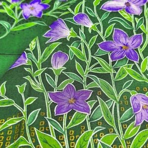 Puede incluir: Primer plano de una tela con un estampado floral. El diseño presenta flores moradas con centros amarillos, hojas y tallos verdes sobre un fondo verde oscuro. Los bordes tienen un borde dorado con pequeñas formas cuadradas.
