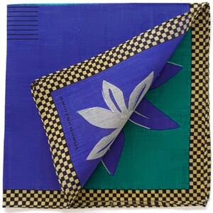 May include: A folded square handkerchief with a blue background and a green section. The handkerchief features a silver floral design and a black and tan checkered border. The text "JUNKO KOSHINO" is printed on the blue section.