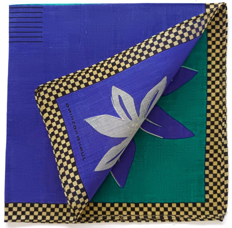 May include: A folded square handkerchief with a blue background and a green section. The handkerchief features a silver floral design and a black and tan checkered border. The text "JUNKO KOSHINO" is printed on the blue section.