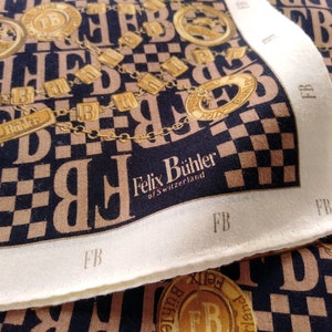 May include: A square silk scarf with a repeating pattern of the letters "FB" in a tan color against a navy blue background. The scarf features gold-colored chain and coin designs, and the text "Felix Bühler of Switzerland".