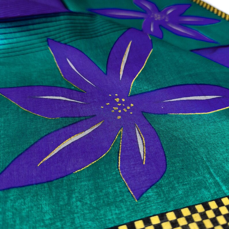 May include: Close-up of a fabric with a floral pattern. Large purple flowers with gold accents are set against a teal green background. The fabric has a black and yellow checkered border.