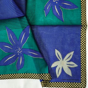 May include: A colorful silk scarf with a floral pattern. The scarf features large blue flowers with gold accents on a teal and blue background. The border has a black and white checkered pattern. The scarf is folded, showing different sections of the design.