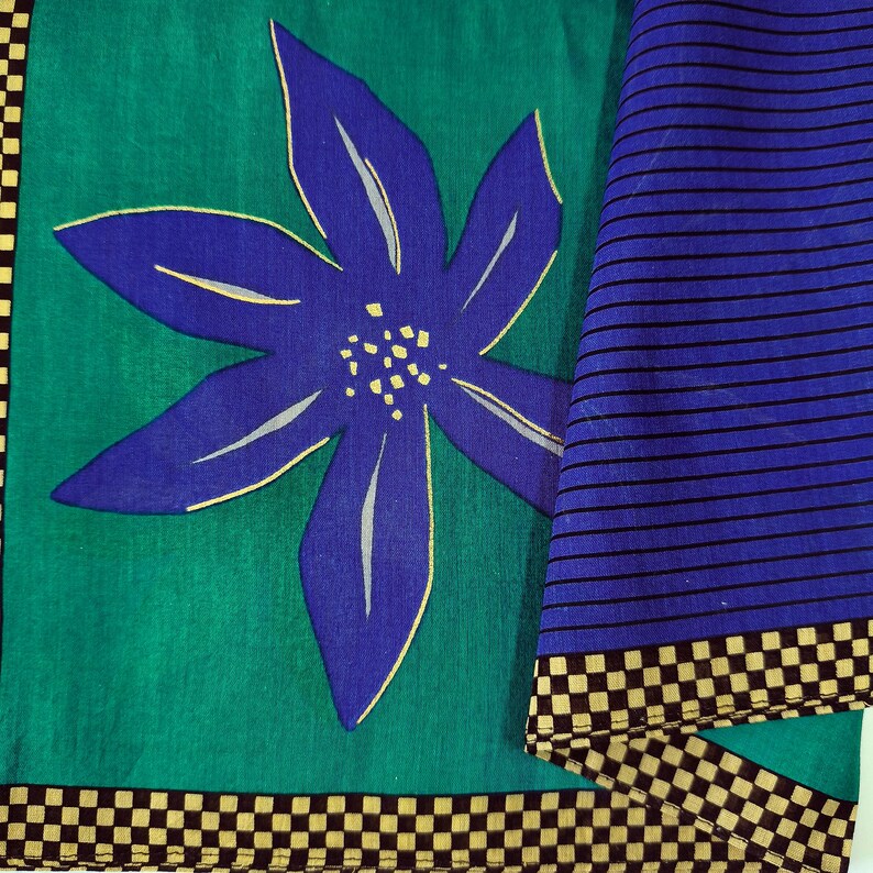 May include: A textile with a large blue flower design on a teal background. The flower has yellow accents. A section of blue fabric with black horizontal stripes is visible. The edges are trimmed with a black and tan checkerboard pattern.