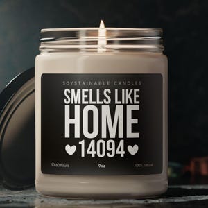 Puede incluir: Una vela de soja encendida en un tarro de cristal con una etiqueta negra. La etiqueta dice "Smells Like Home 14094" en texto blanco. La vela es de 255g y 100% natural. La vela está en un tarro de cristal con tapa plateada.