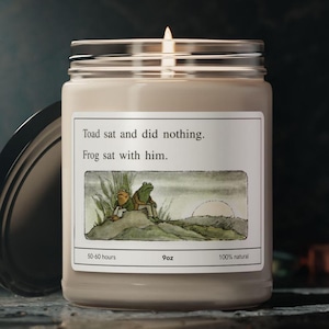 May include: A beige candle jar with a white label featuring an illustration of two frogs sitting on a rock. The label reads "Toad sat and did nothing. Frog sat with him." The candle is 9 ounces and has a burn time of 50-60 hours.