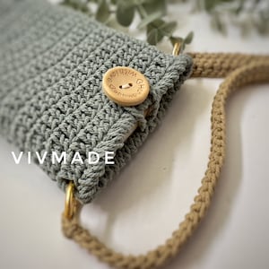 May include: A light green crocheted phone case with a wooden button that says "With Love" in brown lettering. The case is attached to a beige crocheted strap.