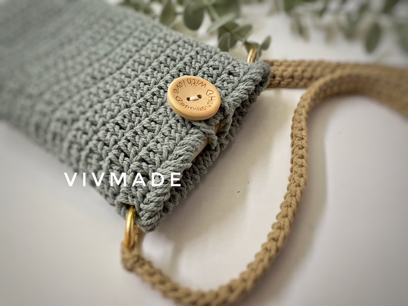 May include: A light green crocheted phone case with a wooden button that says "With Love" in brown lettering. The case is attached to a beige crocheted strap.