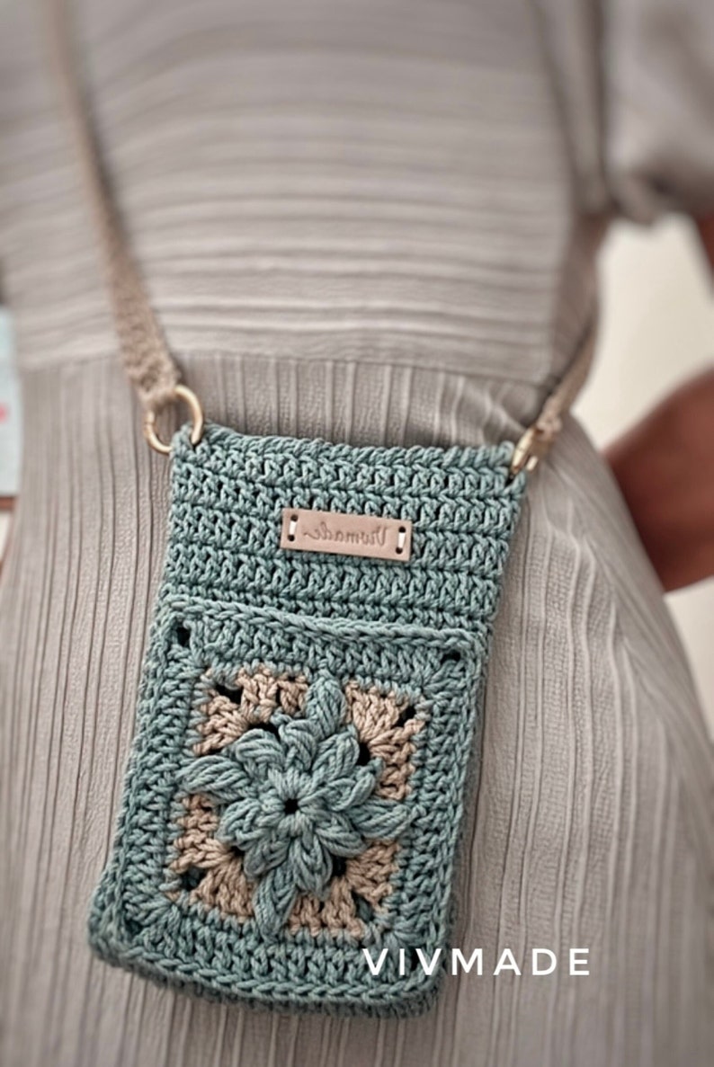 May include: A teal blue crocheted crossbody bag with a brown leather tag that says "remarkable". The bag features a flower design in the centre and is worn over a person's shoulder.