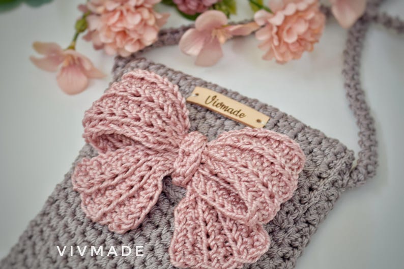 May include: A gray crocheted crossbody bag with a pink crocheted bow and a wooden tag that says "Vivmade".