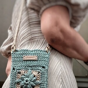 May include: A teal blue crocheted crossbody bag with a brown leather tag that says "Vivmade". The bag features a beige crocheted flower design in the centre.