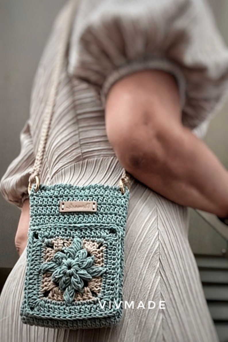 May include: A teal blue crocheted crossbody bag with a brown leather tag that says "Vivmade". The bag features a beige crocheted flower design in the centre.
