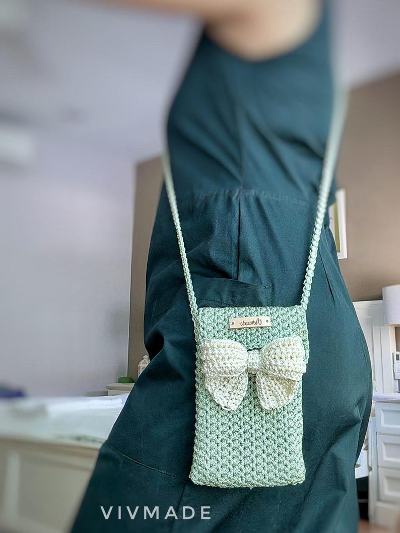 May include: A light green crocheted crossbody bag with a white crocheted bow. The bag has a small label that says "remarkable".