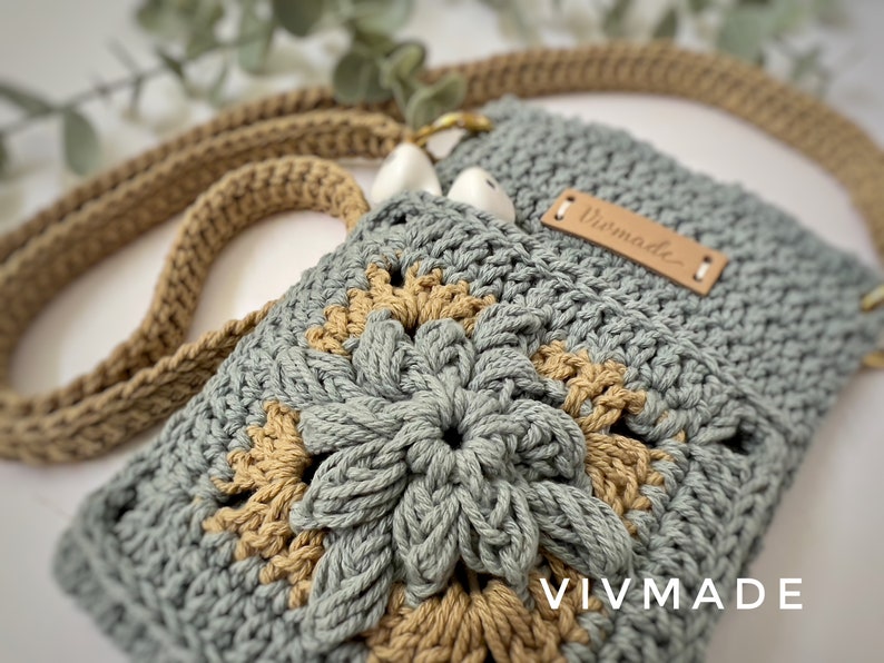 May include: A crocheted bag in a light blue and tan colour scheme. The bag features a flower design and a leather tag that reads "Vivmade".