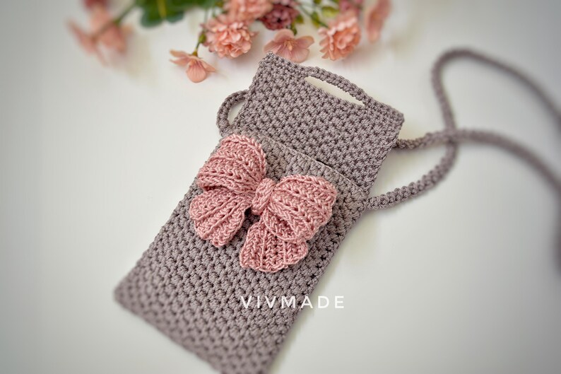 May include: A gray crocheted phone pouch with a pink crocheted bow. The pouch has a strap and the text "VIVMADE" is visible on the front.