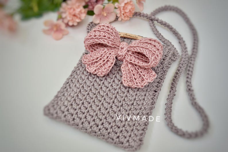 May include: A small crocheted bag with a pink bow and a long strap. The bag has the text "VIVMADE" on the front.
