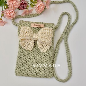 May include: A green crocheted crossbody bag with a cream-colored crocheted bow. The bag has a leather tag with the text "Vivmade" on it.