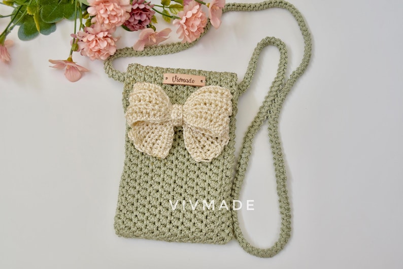 May include: A green crocheted crossbody bag with a cream-colored crocheted bow. The bag has a leather tag with the text "Vivmade" on it.