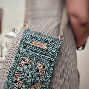 May include: A teal crocheted crossbody bag with a brown leather tag that says "remarkable". The bag features a large crocheted flower in the centre. The bag is being worn by a person.
