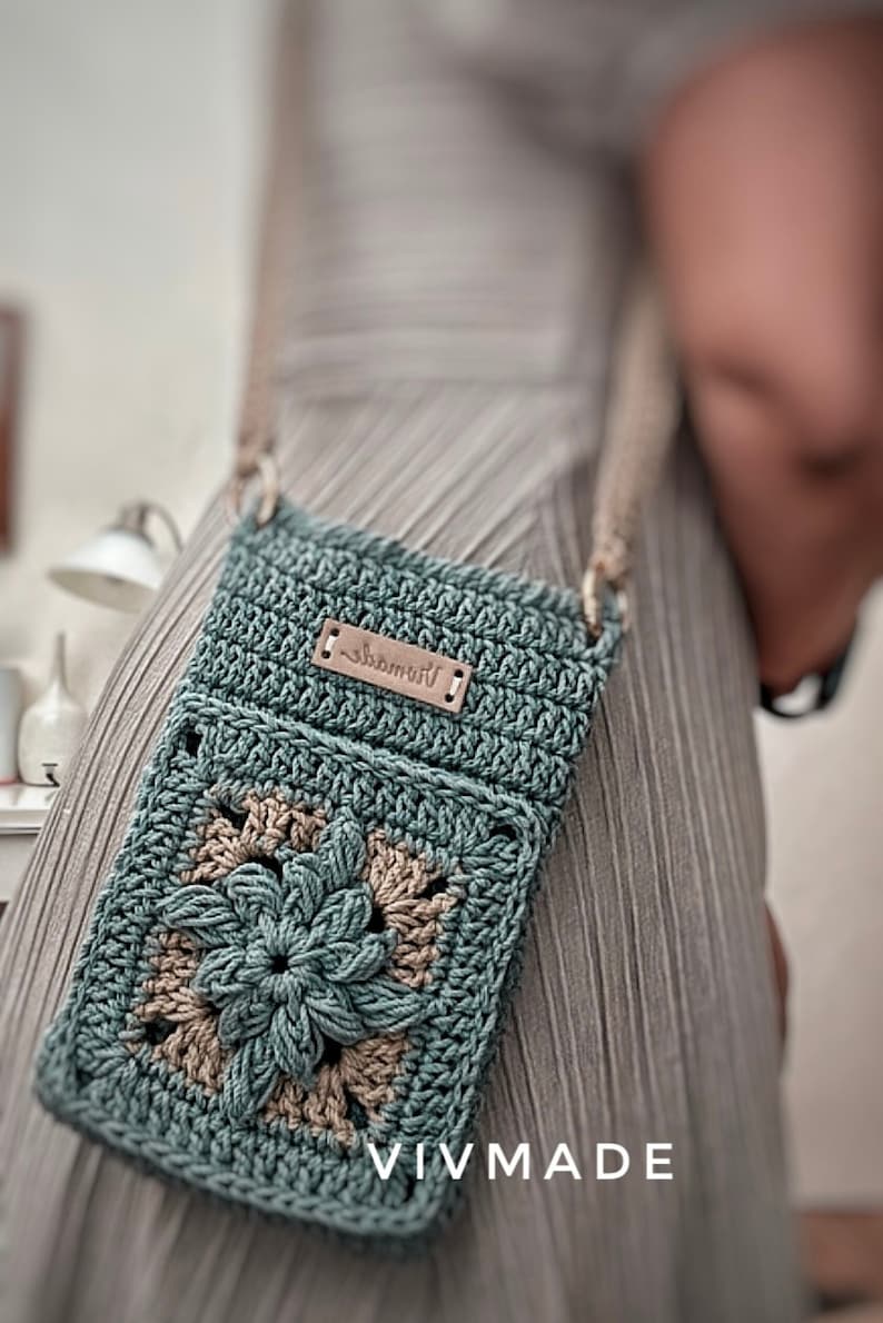 May include: A teal crocheted crossbody bag with a brown leather tag that says "remarkable". The bag features a large crocheted flower in the centre. The bag is being worn by a person.