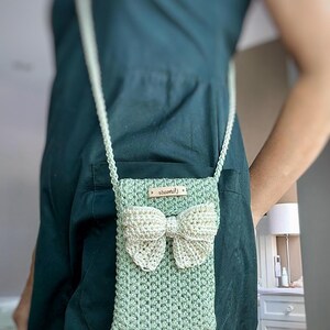 May include: A green crocheted crossbody bag with a white crocheted bow. The bag has a small brown leather tag with the word "handmade" on it.