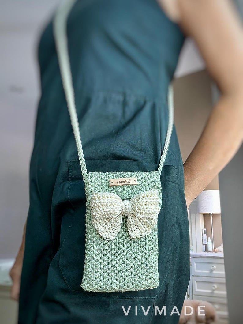 May include: A green crocheted crossbody bag with a white crocheted bow. The bag has a small brown leather tag with the word "handmade" on it.