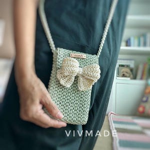 May include: A small green crocheted crossbody bag with a white crocheted bow and a brown leather tag that says "VIVMADE".