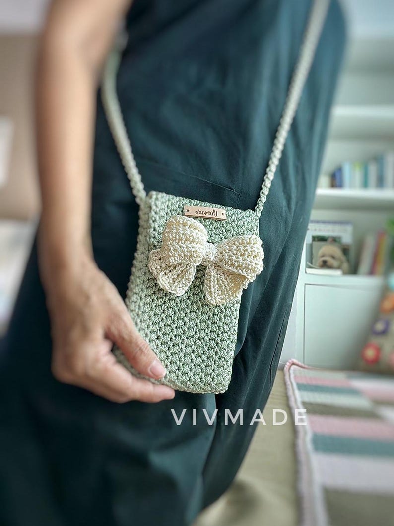 May include: A small green crocheted crossbody bag with a white crocheted bow and a brown leather tag that says "VIVMADE".