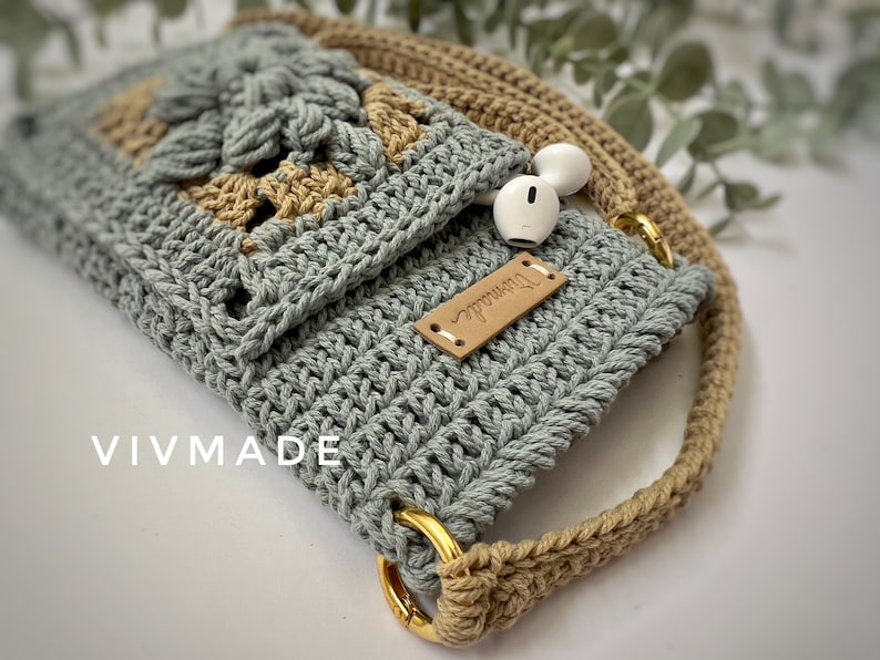 May include: A crocheted phone case with a beige strap and a leather tag that says "remarkable". The case is made with a light blue and beige yarn.
