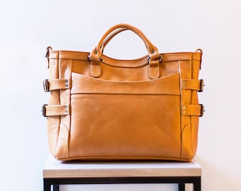 Op de afbeelding: Een bruine leren tote bag met meerdere gespen en banden. De tas staat op een zwarte metalen tafel met een licht houten blad.
