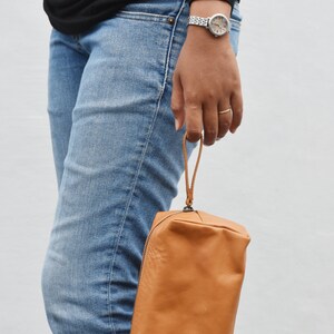 May include: A brown leather zippered pouch with a strap handle. The pouch is being held by a person wearing blue jeans.