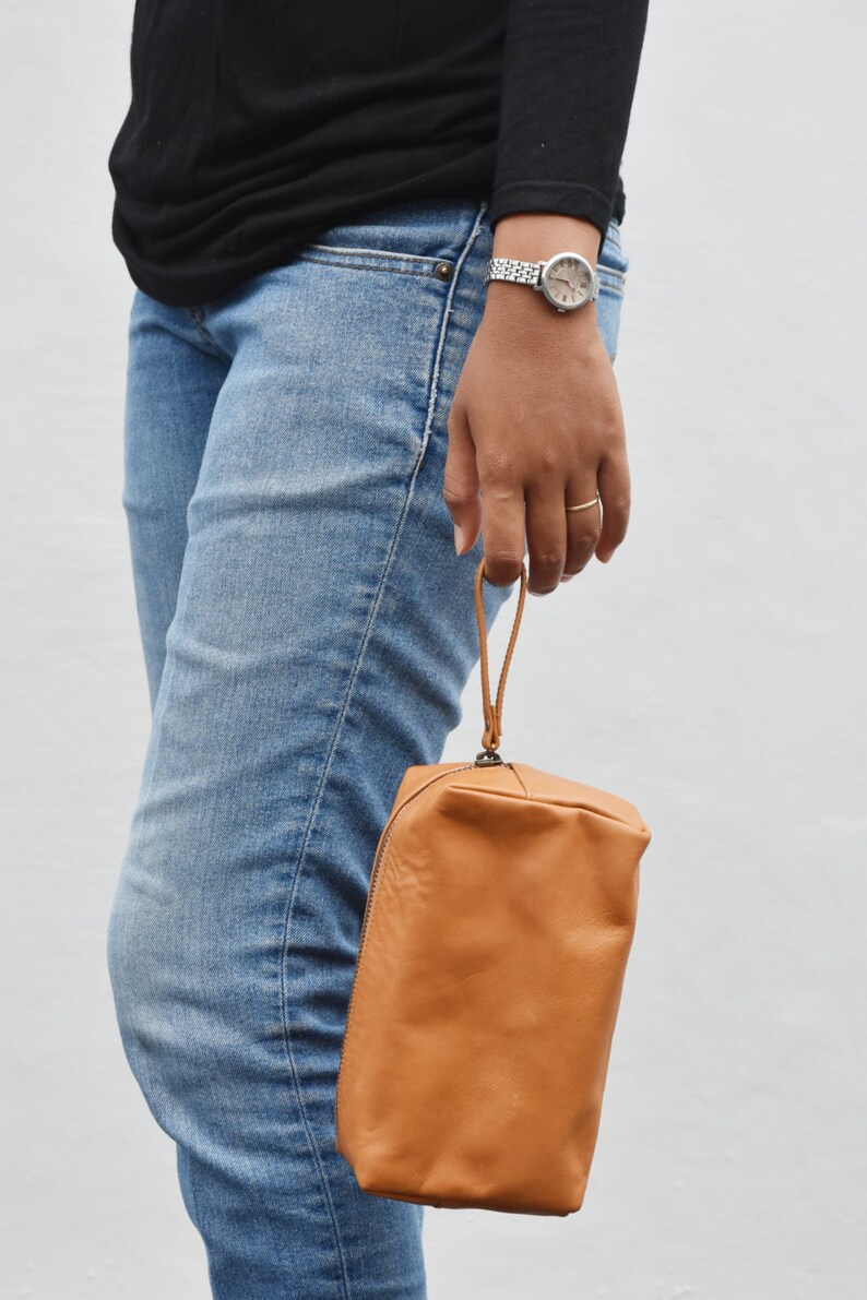 May include: A brown leather zippered pouch with a strap handle. The pouch is being held by a person wearing blue jeans.