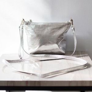May include: A silver leather crossbody bag with a long strap. The bag is sitting on a white table with a black base.