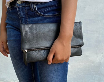 May include: A black leather clutch with a zipper closure and a silver zipper pull. The clutch is held in the hand of a person wearing blue jeans.