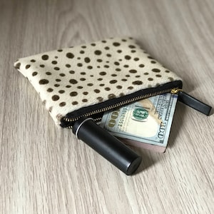 May include: A white and brown spotted zippered pouch with a black zipper pull. The pouch is open and contains a black tube and a folded $100 bill.