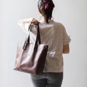 May include: A brown leather tote bag with two straps and gold accents. The bag is being held by a person wearing a white striped shirt and blue jeans.