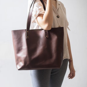 May include: A brown leather tote bag with two straps and gold hardware. The bag is being held by a person wearing a white shirt and gray pants.