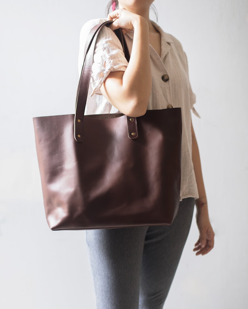 May include: A brown leather tote bag with two straps and gold hardware. The bag is being held by a person wearing a white shirt and gray pants.