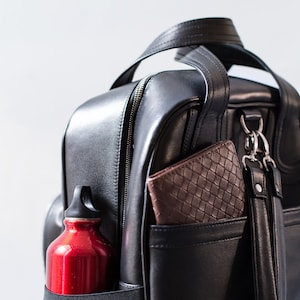 Puede incluir: Una mochila de cuero negro con una botella de agua roja en un bolsillo lateral y una cartera de cuero marr&oacute;n en otro bolsillo.