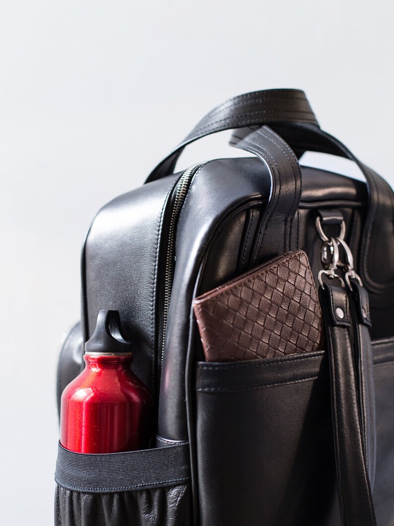 Puede incluir: Una mochila de cuero negro con una botella de agua roja en un bolsillo lateral y una cartera de cuero marr&oacute;n en otro bolsillo.