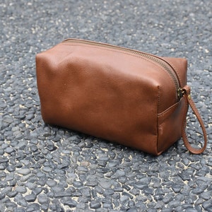 Könnte beinhalten: Eine braune Reise-Toilette-Tasche aus Leder mit Reißverschluss und einem Riemen.