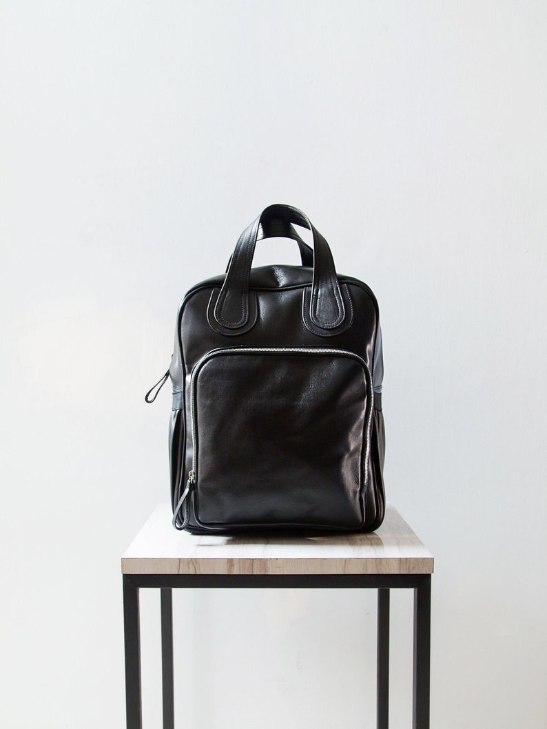 Puede incluir: Una mochila de cuero negro con asa superior y bolsillo frontal con cremallera. La mochila est&aacute; sobre una mesa de madera clara con patas de metal negro.