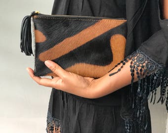 May include: A black and brown cowhide clutch with a zipper closure and a black tassel. The clutch is held in a hand.