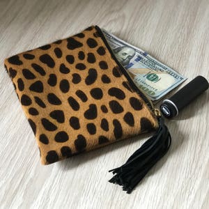 May include: A brown and black leopard print clutch with a black tassel. The clutch has a zipper closure and is open to reveal a dollar bill and a black lipstick tube.