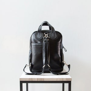 Puede incluir: Una mochila de cuero negro con herrajes plateados y correas ajustables. La mochila est&aacute; sobre una mesa de madera.