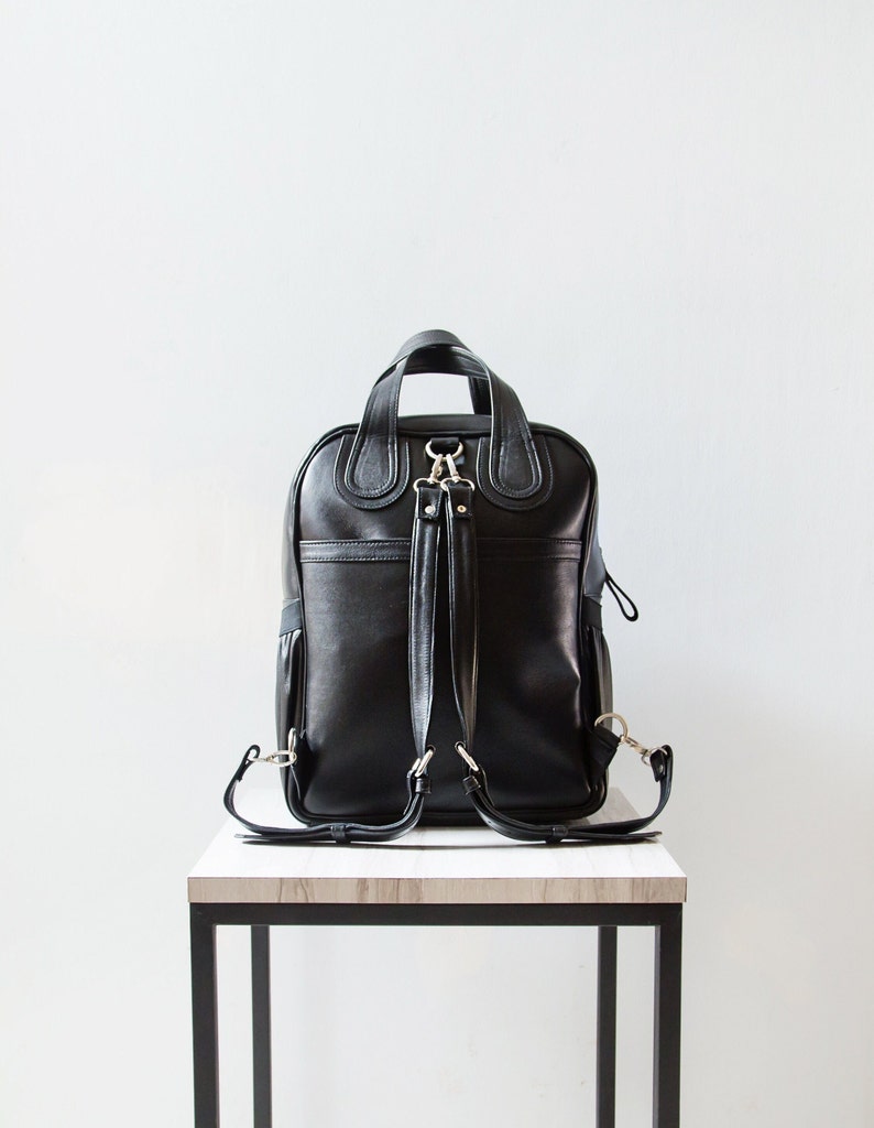 Puede incluir: Una mochila de cuero negro con herrajes plateados y correas ajustables. La mochila est&aacute; sobre una mesa de madera.