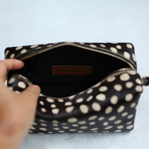 May include: A black and white polka dot makeup bag with a brown leather zipper pull and a gold zipper. The bag is open and the inside is black.