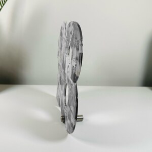 K&ouml;nnte beinhalten: Ein Schwarzwei&szlig;-Fotocollage in Form des Buchstabens "B" ist auf einem klaren Acryl-St&auml;nder montiert. Der St&auml;nder hat eine silberne Metallbasis.